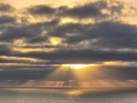 Dramatic sunset over the ocean with golden sunlight breaking through dark clouds, creating beautiful sun rays across the water and a peaceful atmospheric seascape.