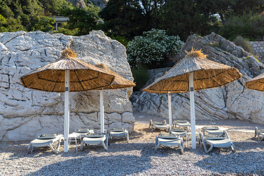 Empty beach loungers under thatched umbrellas in shaded coastal cove with pebble shore, sunlit rocky cliffs, Mediterranean pines, cool blue tones, and calm summer resort atmosphere