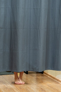 Bare feet visible beneath a fitting room curtain in a clothing store, standing on a wooden floor with soft indoor lighting and ample copy space, illustrating fashion shopping, retail services. 