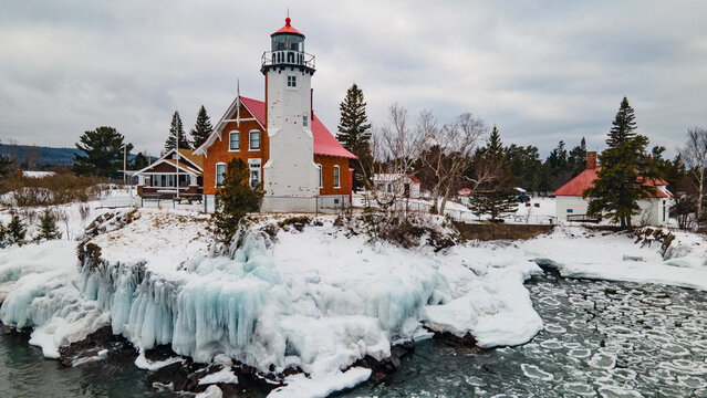 Aerial view of the historic Eagle Harbor Lighthouse surrounded by snow-covered rocky shores and icy waters of Lake Superior in Eagle Harbor, Michigan, United States.