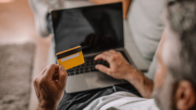 Concentrated Unrecognizable Man Holding Credit Card And Typing On Laptop While Shopping Online At Home. Engaged In Digital Payment Process, Sitting Comfortably In The Living Room Area.