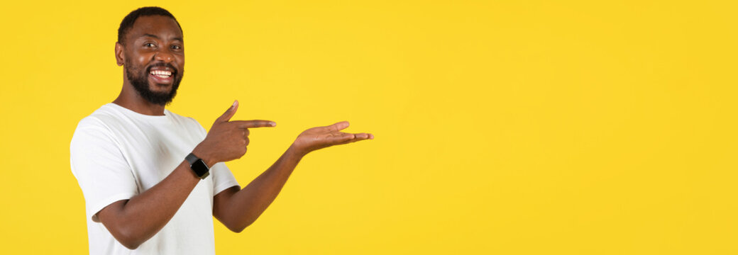 Smiling Black Man In His 30s Pointing At His Open Hand While Standing Against Yellow Background. Ideal For Advertisement Or Marketing Purposes, Showcasing Product Or Service Concept.
