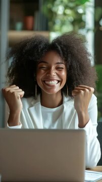 Inspirational of a woman with curly hair smiling and making a fist, expressing accomplishment in an office setting. She is sitting at a desk with a laptop.