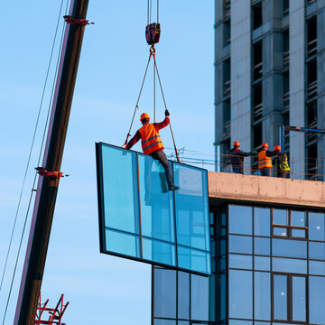Construction workers installing large glass panels on a skyscraper with a crane. The workers are wearing hard hats and safety vests, and the building is still under construction.