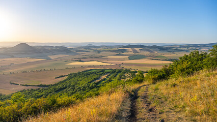 Obraz premium People watch the sunset from Oblik Hill. The sun sets over the fields and valleys in Czechia. The scenery shows rural land with trees and hills under a clear sky.