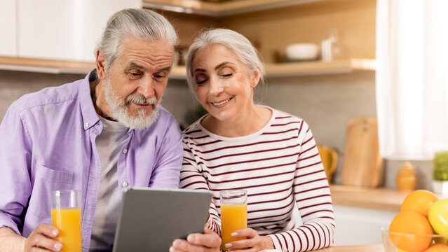 Smiling Elderly Caucasian Man And Woman Sitting Together In Kitchen, Engaged With Tablet And Enjoying Fresh Orange Juice. Happy Couple Sharing Joyful Moments In Their Home Kitchen Setting.
