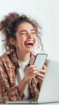 Woman laughs joyfully while holding smartphone. She wears a plaid shirt and uses laptop. Her hair is styled in a loose bun. Background is plain white. Scene conveys happiness and digital engagement