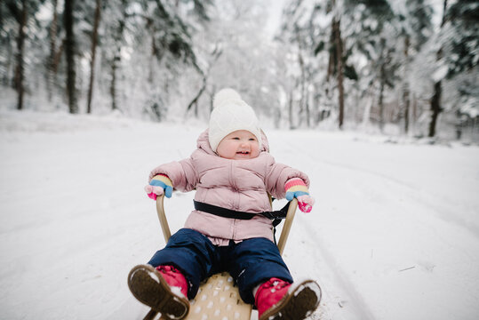 Happy child in winter clothes sitting on sled during walk through snowy forest. Joyful kid mood in winter time. Closeup.