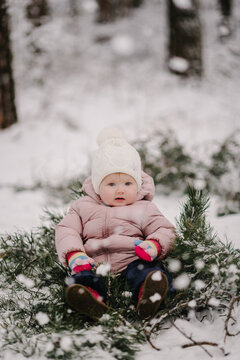Cozy winter portrait of a toddler: child in warm puffer jacket sitting on spruce branches during snowfall. Closeup.