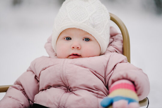 Portrait of a cute little baby girl sitting in a sled in a snowy winter forest, wearing winter clothes. Child looking in camera. Closeup kid.