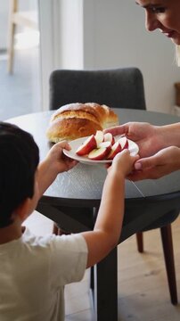 Jewish child carries sliced apples to a Rosh Hashanah table as an adult family member receives the plate, with round challah and honey visible in soft natural daylight.