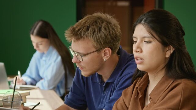 Caucasian young adult male student with glasses explaining project to Latin female classmate in university classroom. Ideal for higher education marketing and student collaboration concepts
