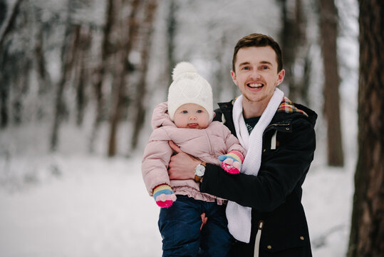 Father gently holding little daughter in his arms during winter walk in snowy forest. Both smiling cheerfully. Cozy family moment in nature. Close up.