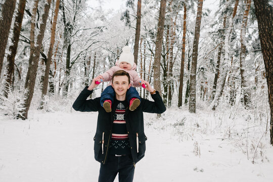 Smiling father carrying his little daughter on shoulders during a walk in the snowy forest. Winter family vacation, happy childhood in nature. Closeup.