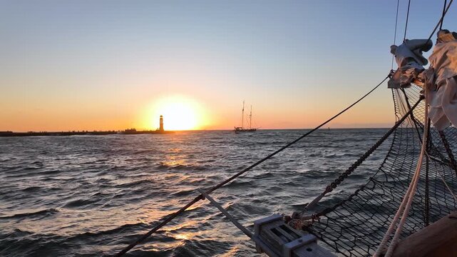 Sailboat bow with net rigging pointing directly at a bright setting sun and lighthouse silhouette on a wavy sea horizon.