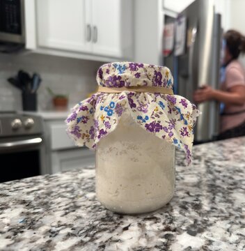 Baker weighing, feeding, discarding active sourdough starter in glass mason jars on a granite kitchen counter, preparing wild yeast for artisan baking