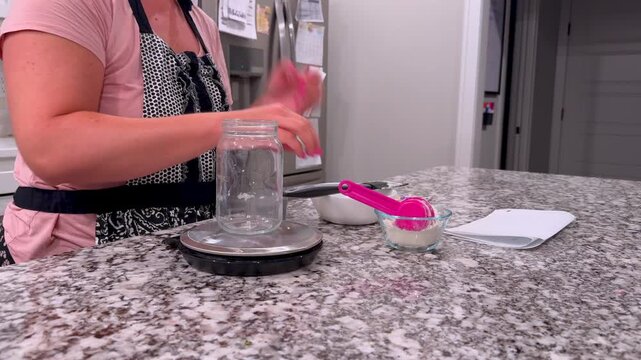 Baker weighing, feeding, discarding active sourdough starter in glass mason jars on a granite kitchen counter, preparing wild yeast for artisan baking