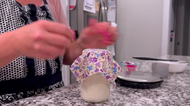 Baker weighing, feeding, discarding active sourdough starter in glass mason jars on a granite kitchen counter, preparing wild yeast for artisan baking