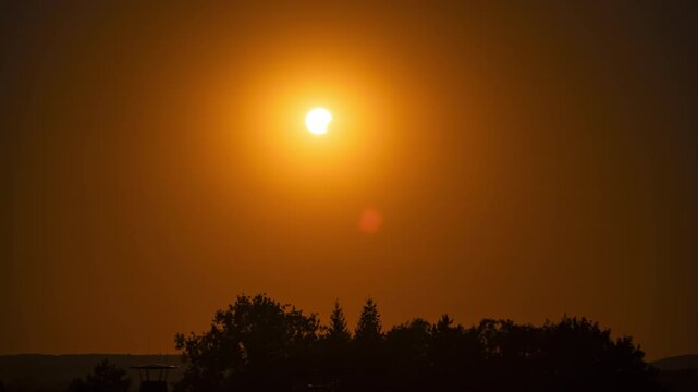 Timelapse of a partial solar eclipse during sunset in the Czech Republic, showing the partially obscured Sun descending toward the horizon behind silhouetted trees.