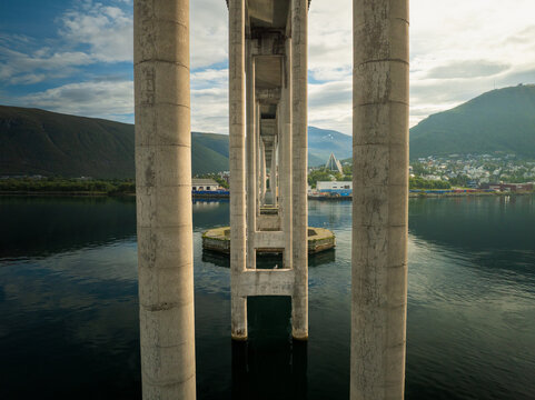 Aerial view of the Tromso Bridge structure with concrete pillars framing the Arctic Cathedral and the coastal town of Tromso, Troms, Norway.