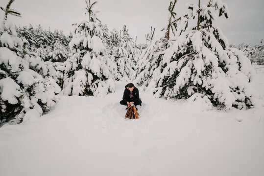 A man warming himself by a small bonfire in a snowy winter forest. A cozy moment outdoors among snow-covered pine trees.