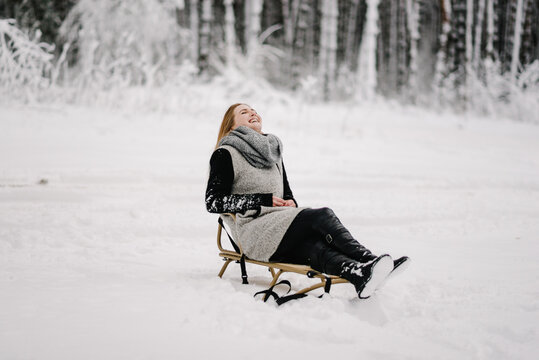 Young woman laughing happily while sitting on a sled in the middle of a snowy winter forest. She is enjoying a bright moment of winter leisure outdoors.