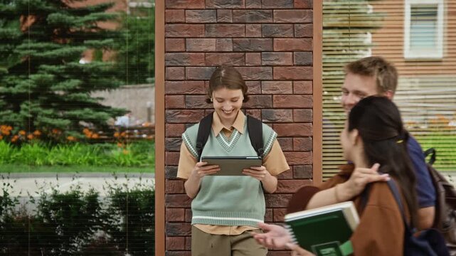 Caucasian teenager student leaning against brick pillar, using tablet on campus while diverse students walk by. Footage suitable for promoting e-learning apps and modern university life