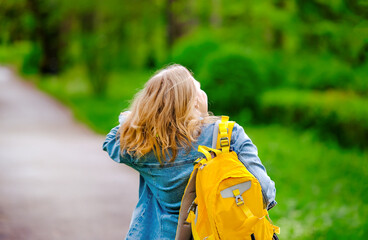 woman with yellow backpack walks away along path in sunny green park. Theme of peaceful solo travel and nature exploration © 5ph