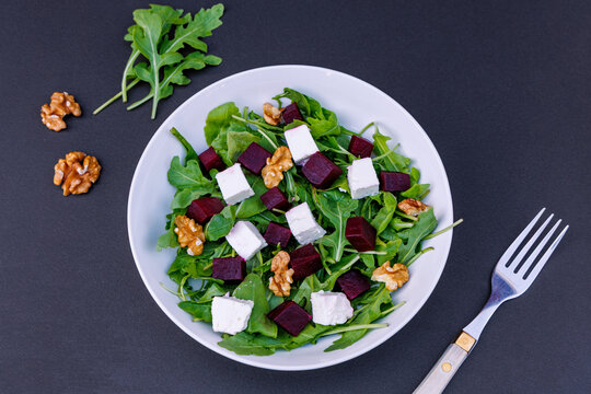Top view of beetroot salad with feta cheese, fresh arugula and walnuts in white bowl on dark background, healthy eating and vegetarian diet concept