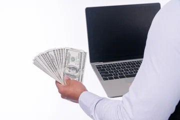 Close up of hand holding a fan of hundred US dollar bills and a laptop computer isolated on white background with copy space. Making money online, e-commerce, and financial success concept. © kaew6566