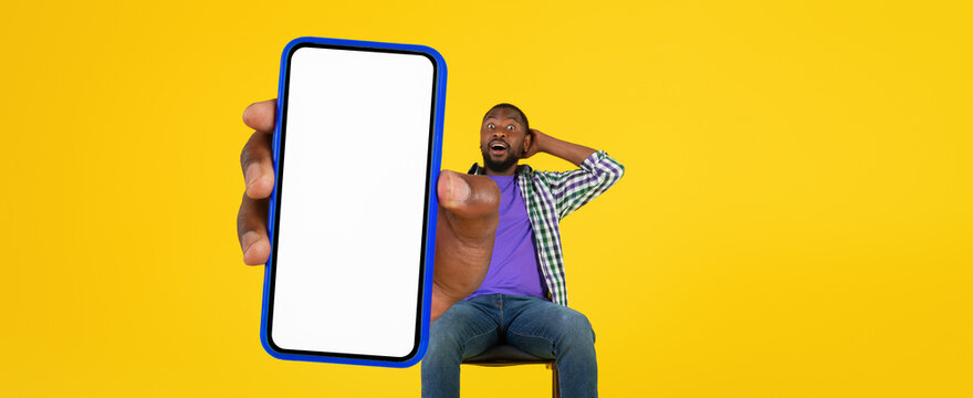 Surprised Black Man Holding Smartphone With Blank Screen, Emotion Of Shock On His Face, Sitting In A Studio Environment With A Yellow Background, Concept Of Technology And Modern Lifestyle.