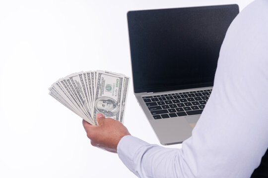 Close up of hand holding a fan of hundred US dollar bills and a laptop computer isolated on white background with copy space. Making money online, e-commerce, and financial success concept.