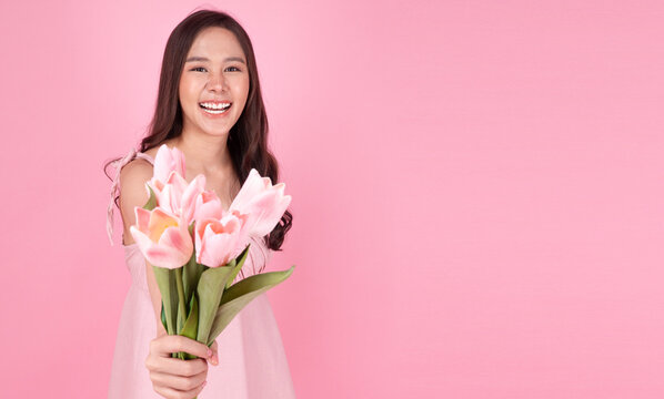 Beautiful smiling young Asian woman in pink dress holding and giving a bouquet of pink tulip flowers isolated on pink background with copy space. Valentine, Mother's day, and celebration concept.