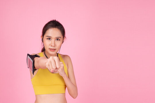 Portrait of a determined young Asian woman in yellow sportswear punching toward camera on pink background with copy space. Female fitness workout and martial arts training concept.