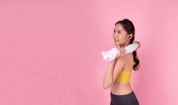 Portrait of happy young Asian woman in yellow sportswear with white towel around neck looking away at copy space on pink background. Female fitness, workout and active lifestyle concept.