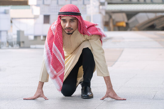 Portrait of an Arab businessman in traditional clothing getting ready in a runner's starting position on a city street. Business competition, startup, and race to success concept.