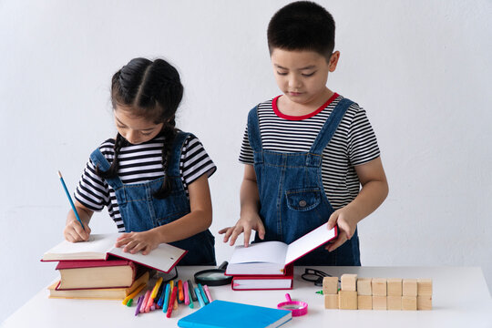 Two cute young Asian children, boy and girl in matching outfits, reading and writing on books at a desk isolated on white background with copy space. Sibling education and childhood learning concept.