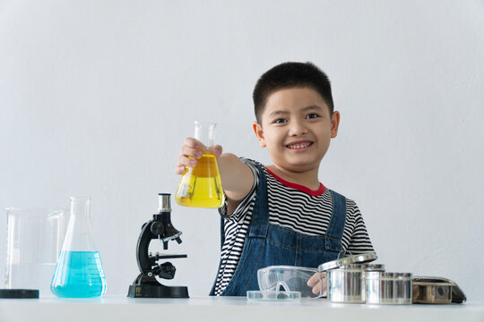 Smiling young Asian boy holding a chemistry flask with yellow liquid. Little student doing science experiment with microscope on desk isolated on white background. STEM education concept.
