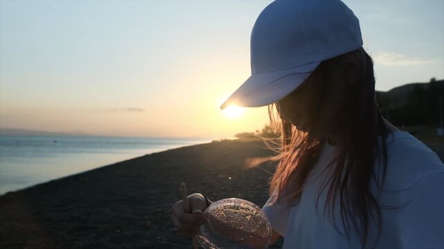 Preteen girl in a white cap at sunset on the coast opening a container of hot instant noodles, removing a round plastic lid covered with condensation, ready for an outdoor hot meal