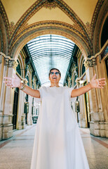 Obraz premium Woman in white dress with sunglasses stands in grand architectural corridor, arms outstretched, showcasing ornate arches and glass ceiling in a historic building
