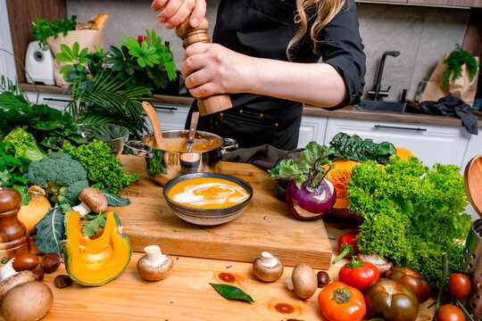 Woman with pepper mill over bowl of pumpkin soup on kitchen table with fresh vegetables. Gourmet spice seasoning concept for autumn recipe, healthy organic dinner, and culinary presentation.
