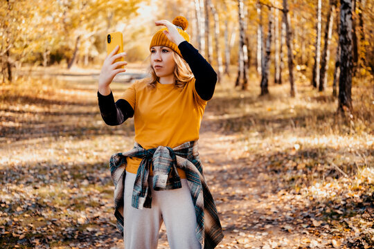 Female tourist in yellow beanie taking selfie with smartphone while hiking in autumn forest. Modern digital communication and social media sharing.