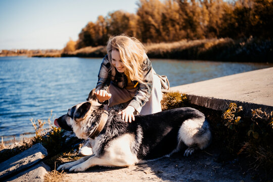Woman pets Shepherd dog near river in outdoor autumn park. Happy domestic animal friendship concept.