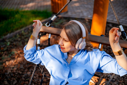 Woman in headphones relaxing in outdoor park wooden hammock chair. Calm modern outdoor leisure concept.