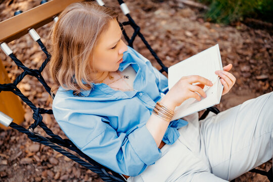 Woman in blue shirt reads book relaxing in rope hammock. Leisure time outdoors on sunny summer day.