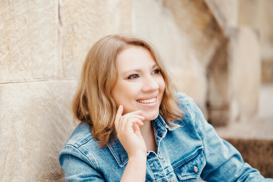 Happy woman in denim jacket sits near stone wall smiling looking away in city. Concept of positive emotions and urban leisure time.