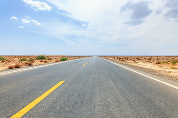 Straight paved highway crossing the arid desert landscape of Xinjiang, China, with sparse shrubs, yellow centerline, and expansive blue sky for travel and infrastructure themes. © zhao dongfang