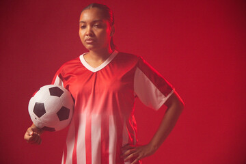 Obraz premium Adult African woman standing in red studio wearing red-white jersey holding black-white ball