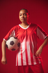 Obraz premium African American woman posing in studio wearing red-white kit, holding black-white ball at hip