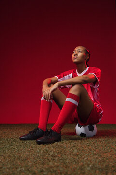 African-American female soccer player sitting on ball on studio turf, gazing up in red kit, cleats